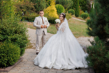 A bride and groom are standing in a garden, the bride holding a bouquet. The bride is wearing a long white gown and the groom is wearing a suit. The scene is romantic and picturesque