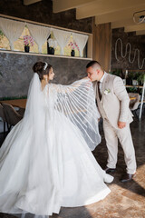 A bride and groom are standing in a room with a white curtain behind them. The bride is wearing a white dress and a veil, while the groom is wearing a suit. They are both smiling