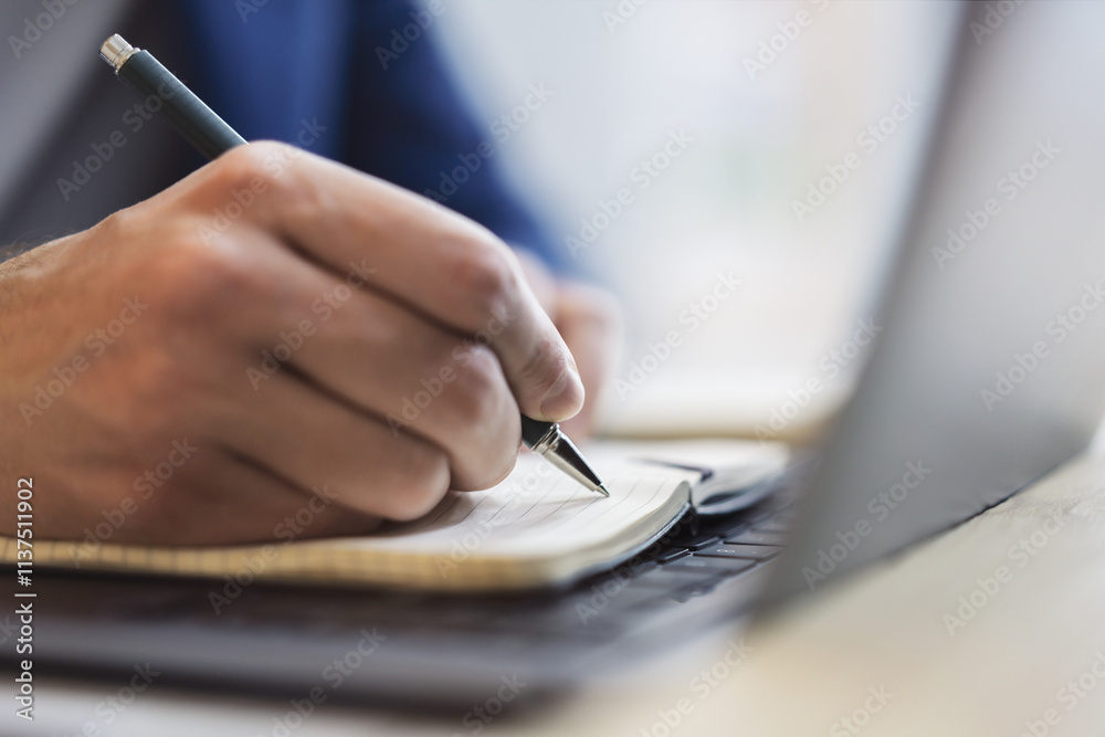 Poster detailed view of a man's hand jotting down notes in a notepad on a modern laptop, with a blurred bac