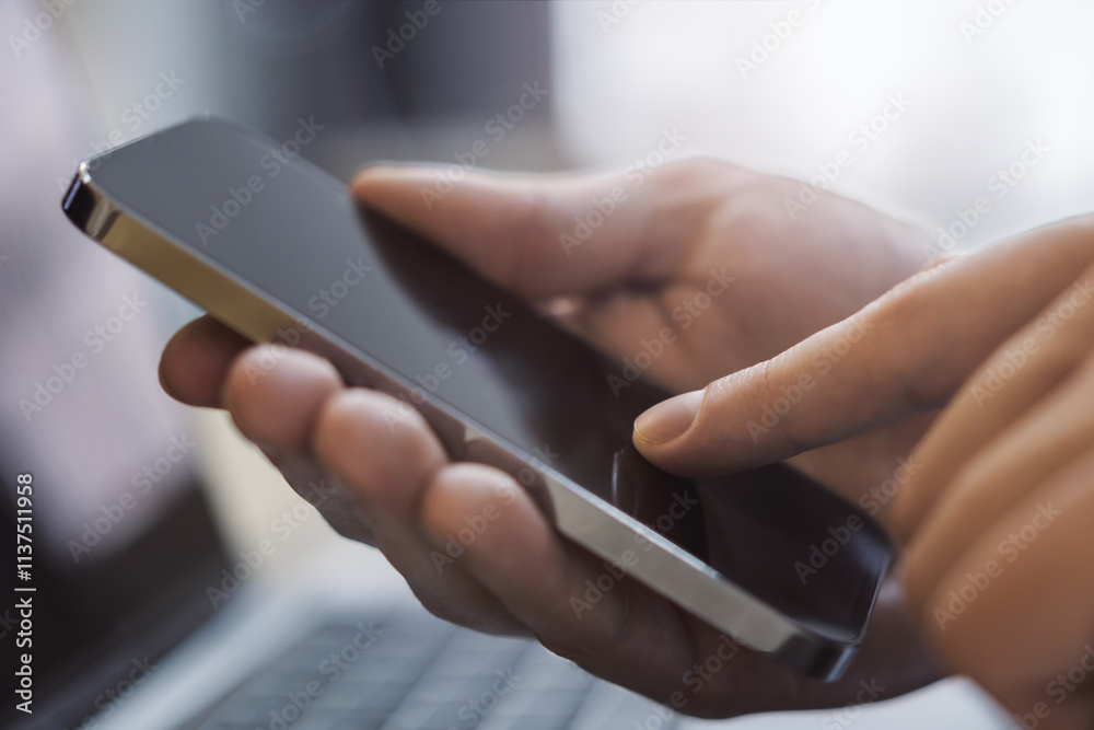 Poster closeup view of a man engrossed in his smartphone tasks with an open laptop nearby set against the b