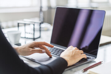 The image shows a woman's hands in sharp focus, typing on a modern laptop keyboard, office scene blurred in the backdrop