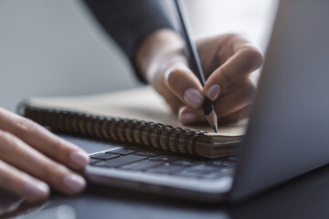 Detailed view of a woman's hand taking notes in a notepad on a cutting-edge laptop, against a...