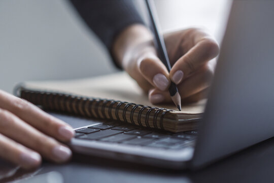 Detailed view of a woman's hand taking notes in a notepad on a cutting-edge laptop, against a blurred backdrop