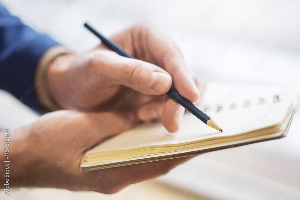 Poster focused view of a man's hand as he writes in a notepad, background gently blurred