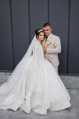 Fototapeta premium A bride and groom are posing for a picture in front of a gray wall. The bride is wearing a long white gown with a veil and a tiara, while the groom is wearing a suit