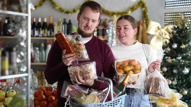 Couple buys delicious products in the Christmas supermarket. Man and woman with sweets and tangerines next to food shelves