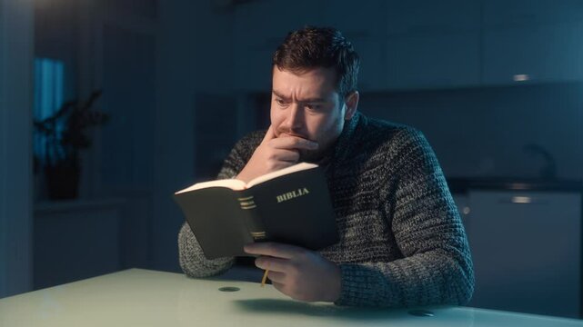 Bearded man reflecting on faith while reading the Bible in a darkened room.