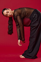 Fashion asian woman in black pants and sheer top bending over on red background with hands on knees...