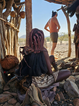 Himba woman with traditional otjize hairstyle sitting inside a hut made of wooden poles, interacting with a tourist who is observing the village life