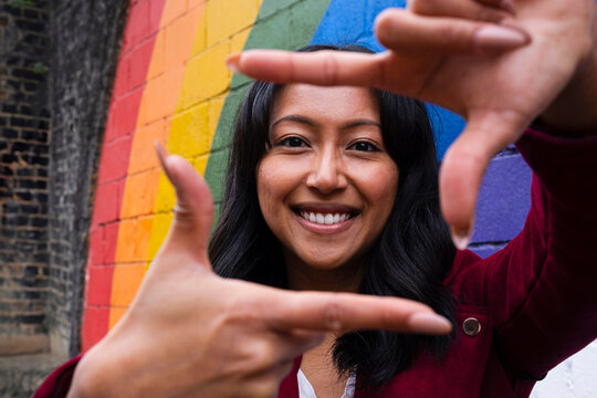 Smiling woman looking through finger frame in front of rainbow wall