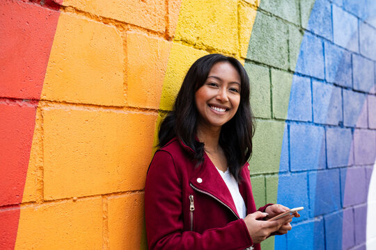 Smiling woman leaning with smart phone on rainbow wall