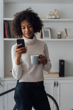 Mid adult freelancer standing in her home office, using smartphone, holding coffee cup
