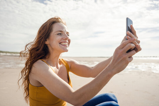 Netherlands, Zeeland, grinning redheaded woman taking selfie with smartphone on the beach