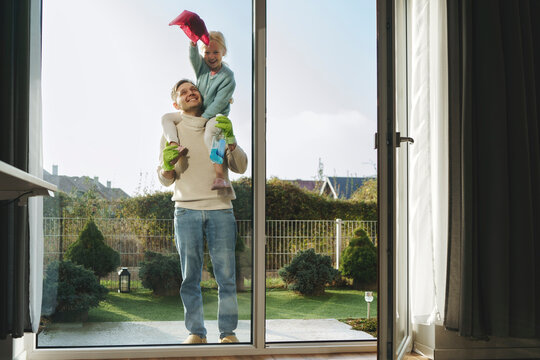 Smiling father carrying daughter on shoulders and cleaning window