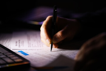 Close-up of Hand Writing on Paper with Black Pen Next to Laptop in Dim Lighting