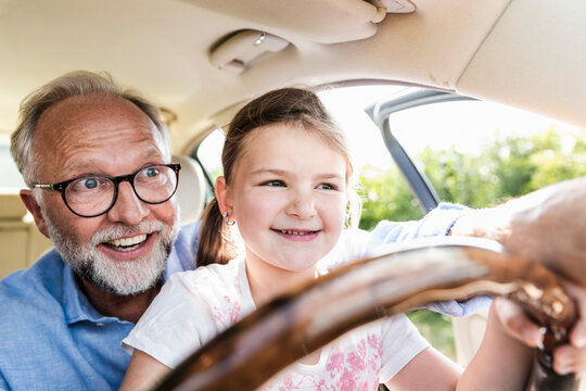 Little girl sitting on lap of grandfather, pretending to steer the car