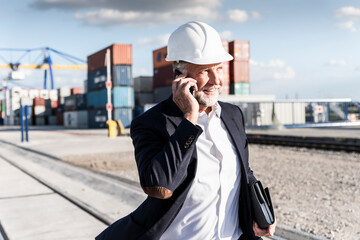 Businessman at cargo harbour, wearing safety helmet, using smartphone