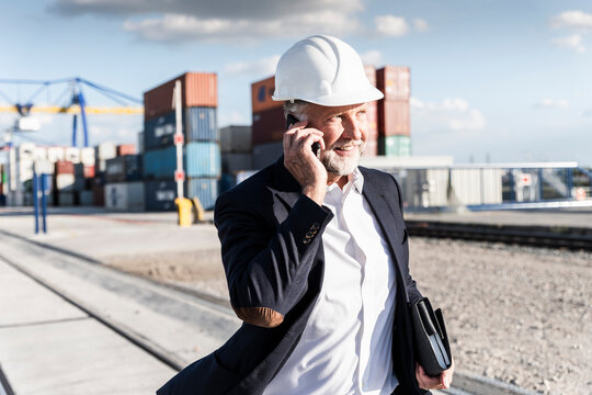Businessman at cargo harbour, wearing safety helmet, using smartphone
