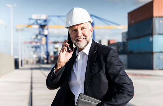 Businessman at cargo harbour, wearing safety helmet, using smartphone