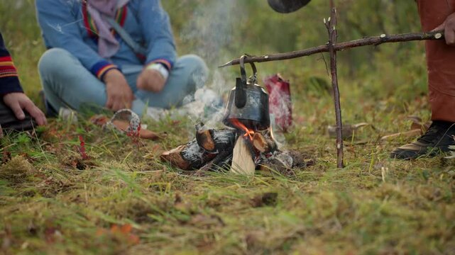 Low wide view of metal kettle boiling on wood flame, Sami family brewing coffee over an open fire in Narvik Norway surrounded by lush greenery