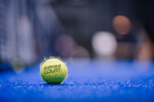  Close-up of a yellow Babolat padel ball on a blue court surface, with a blurred background in an indoor sports facility. Padel.
