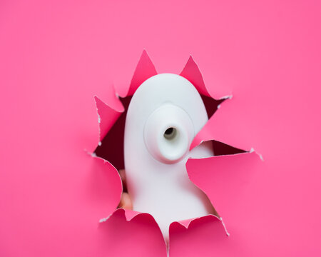 White vacuum clitoral vibrator sticking out through pink paper background. Sex toy. 