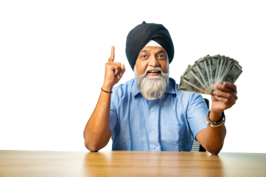 Elderly Sikh man with Indian currency fan, sitting at table, showing happy expression, success sign