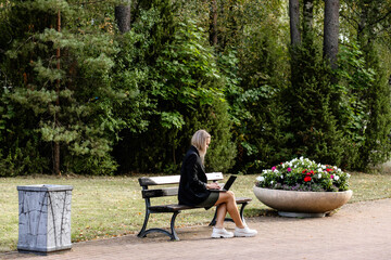 Businesswoman sitting on bench in summer park using, typing on laptop computer, studying outdoors. Female blond person working remote. 