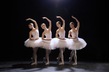 Four young and beautiful ballerinas in white tutus showing perfect symmetry during graceful performance eon dimly lit stage on black background. Concept of classical dance, art, beauty of movements