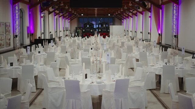 Elegant banquet hall with white table settings, candles, and covered chairs, featuring a railway theme with vintage trains in the background under soft purple lighting.