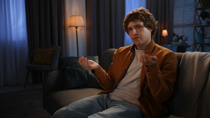 Fototapeta premium Young man spending evening at home sitting on sofa in living room, holding coins in hands looking upset at camera, disappointed hopeless expression.