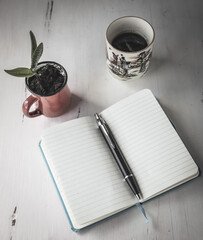 Foto de un cuaderno a rayas en blanco abierto sobre un escritorio junto a una pequeña planta y una taza de cafe.