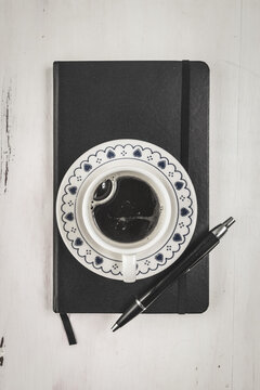 Foto cenital de una taza de cafe negro sobre un cuaderno de notas cerrado junto a una lapicera sobre una mesa blanca