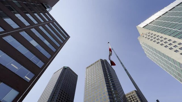 Red and white Canadian flag flutters atop a flagpole set against a backdrop of modern skyscrapers, with a clear blue sky emphasizing the urban scene, Canada