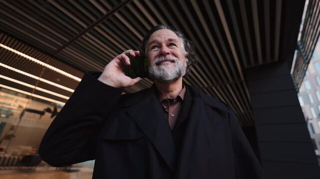mature bearded man engages in mobile phone call while walking near modern building. Male entrepreneur focused and attentive conversating with smartphone device, enjoying communication technology 
