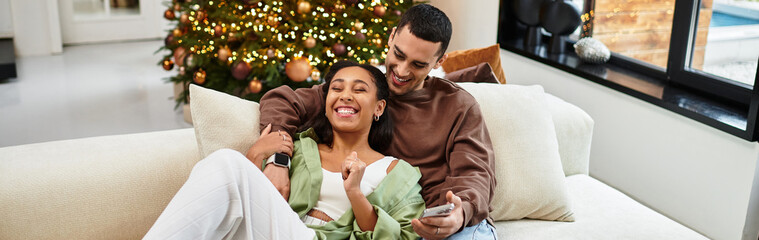 Fototapeta premium Joyful couple relaxes together on a sofa, surrounded by Christmas decorations and lights, banner