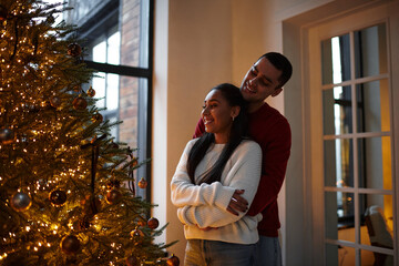 A beautiful couple shares a warm embrace by the shimmering Christmas tree, celebrating love.