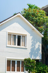 A white, wood-sided house with a sloped roof and blue sky, a leafy, green tree with red flowers grows over the roof