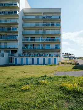A modern building near Hardelot features balconies and blue storage doors, surrounded by green and yellow wildflowers
