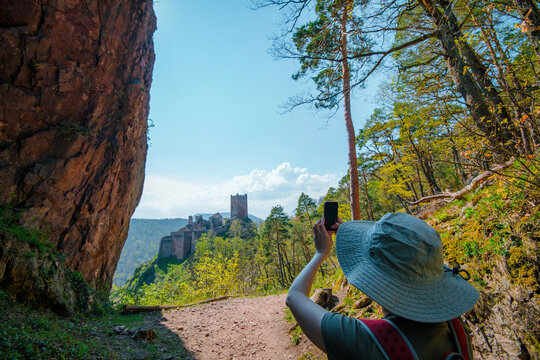 A woman capturing the view of Chateau de Fleckenstein in Alsace, surrounded by dense greenery and rocky landscapes under a bright blue sky