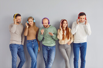 Portrait of a young joyful happy people friends or students men and women wearing casual clothes having fun listening to music in headphones standing in a row on a gray wall background.