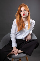 Portrait of a red-haired girl in a white shirt. Woman sitting on a chair in the studio against a gray background.