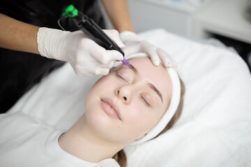 Skilled hands apply permanent makeup to a relaxed woman lying comfortably in a beauty salon. The procedure focuses on enhancing her eyebrows with precision and care.