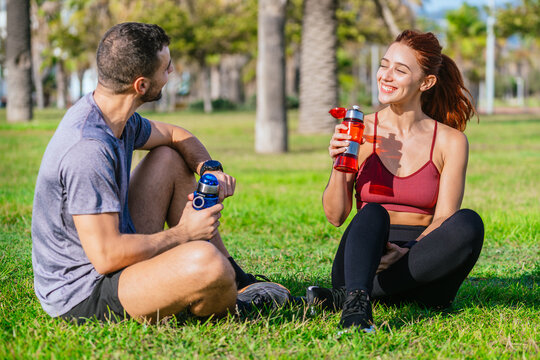 A fit man and woman sit on the grass in a sunny park, smiling and holding water bottles. They are dressed in sporty attire, relaxing and staying hydrated after a workout session surrounded by trees