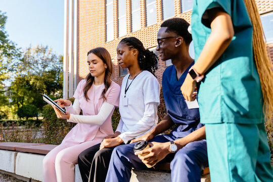 Multicultural nursing students in different colors scrubs are happily interacting and sharing notes outside medical facility.