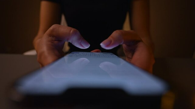 Top view of unrecognizable woman using typing smartphone in dark room, browsing online content with illuminated screen. Female searching web while reading news and blog posts online, slow motion.