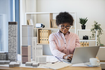 African woman architect working on laptop to design modern city model in office setting. Professional, focused, middle-aged woman with architectural expertise and creativity.