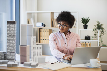 African woman architect in office designing model of modern city with laptop and skyscrapers. Young female professional in architecture field focused on project.