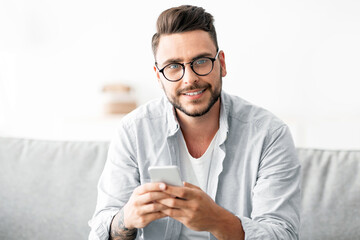 Positive young guy with glasses sitting on couch, using smartphone and smiling to camera, chatting with friends and chilling at home, copy space