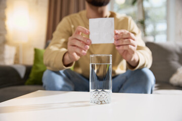 Close up of young sick man shows white sachet, preparing to take effervescent tablet while sitting at table on sofa in living room. Concept of illness, healthy insurance, pharmaceutical, medicine. Ad.